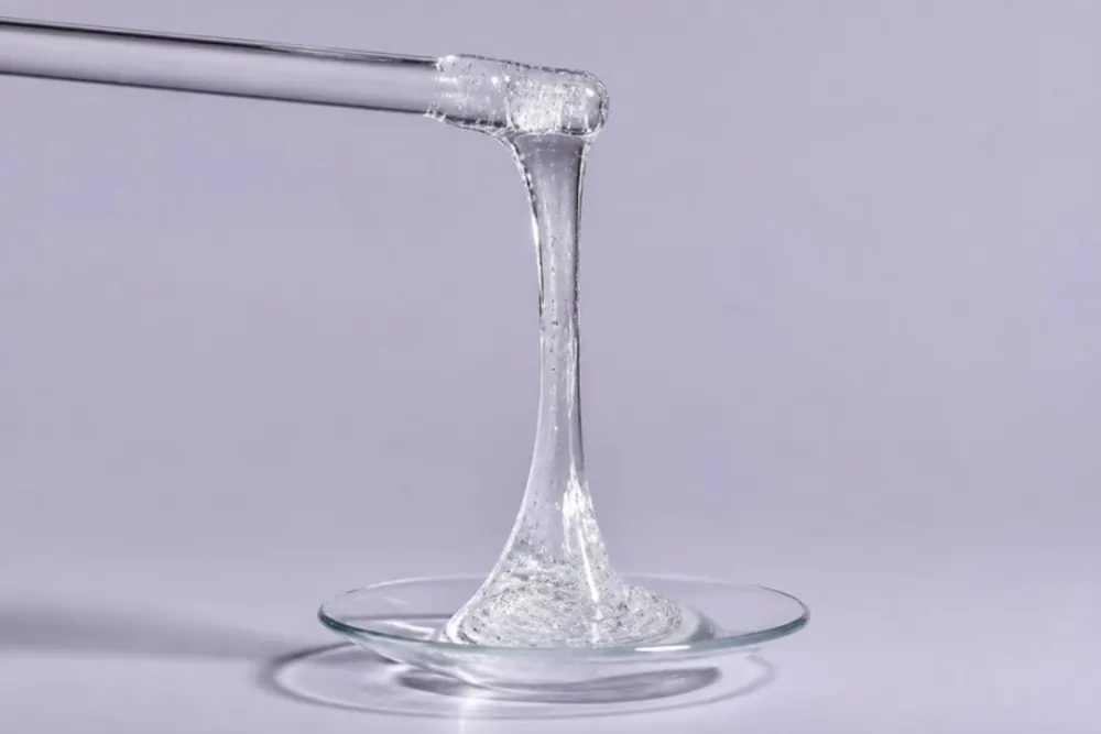 Gel transparente y viscoso que se estira en un hilo entre una varilla de vidrio y un vidrio de reloj, sobre un fondo lila grisáceo.