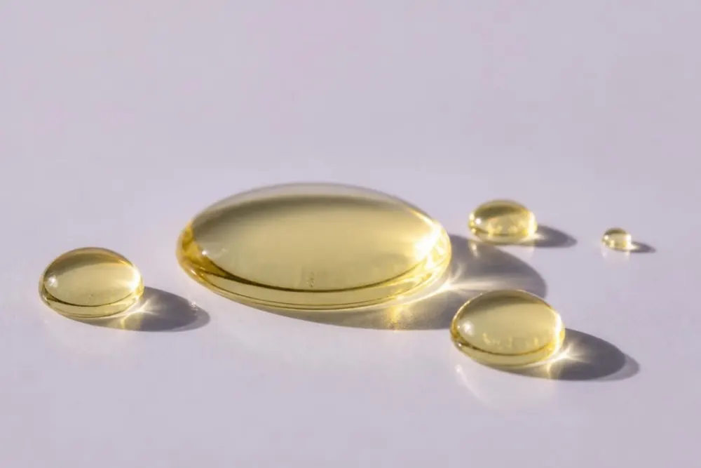 Gotas abombadas de aceite dorado pálido sobre una lámina de vidrio, con fondo lila grisáceo