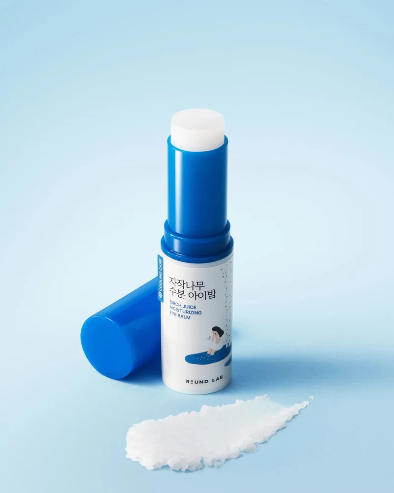 Barra de Birch Moisturizing Eye Balm abierta, con la tapa al lado, sobre fondo azul